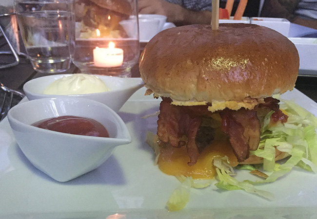 Mit Cheddar überbackener Prime Burger aus 100% Black Angus Beef, serviert im gebutterten Weizenbun mit crossem Speck, Eisbergsalat und Tomato– Relish (22,80 EUR)